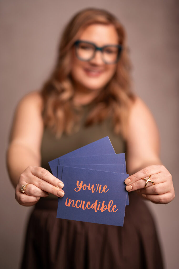 Kristine Depies, founder of KAYDEE Management Solutions, holding a stack of navy blue cards that read “You’re incredible” in orange script. Part of the Power of Her headshot series by Kara Reese Photography.