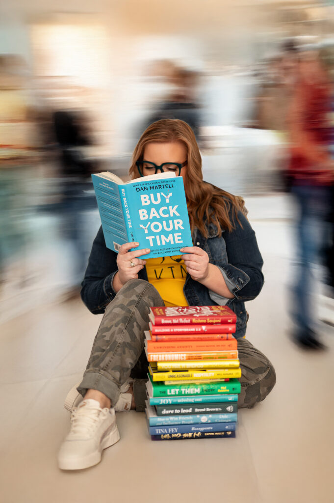 Kristine Depies, founder of KAYDEE Management Solutions in Waukesha, WI, sits on the floor reading “Buy Back Your Time” by Dan Martell, surrounded by a colorful stack of business books. Blurred crowd movement creates a sense of calm amidst chaos. Branding photo by Kara Reese Photography for the Power of Her campaign.